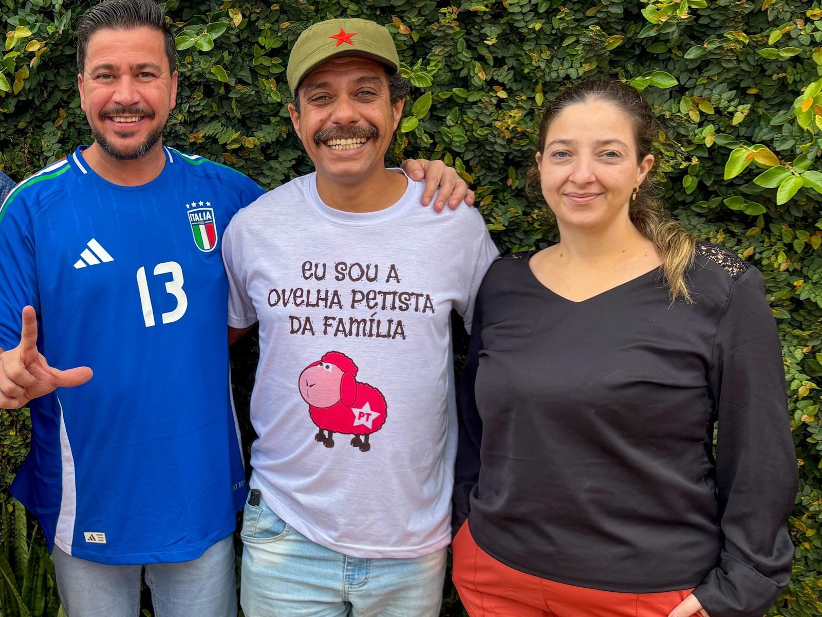 O deputado Arilson Chiorato (esquerda), o jornalista Tiago Prado (centro) e sua esposa, a palestina Amani Said (direita), posam juntos. Tiago veste uma camiseta com os dizeres "Eu sou a ovelha petista da família" e um boné verde com estrela vermelha, simbolizando o apoio político e a união da militância de esquerda no PT do Paraná.