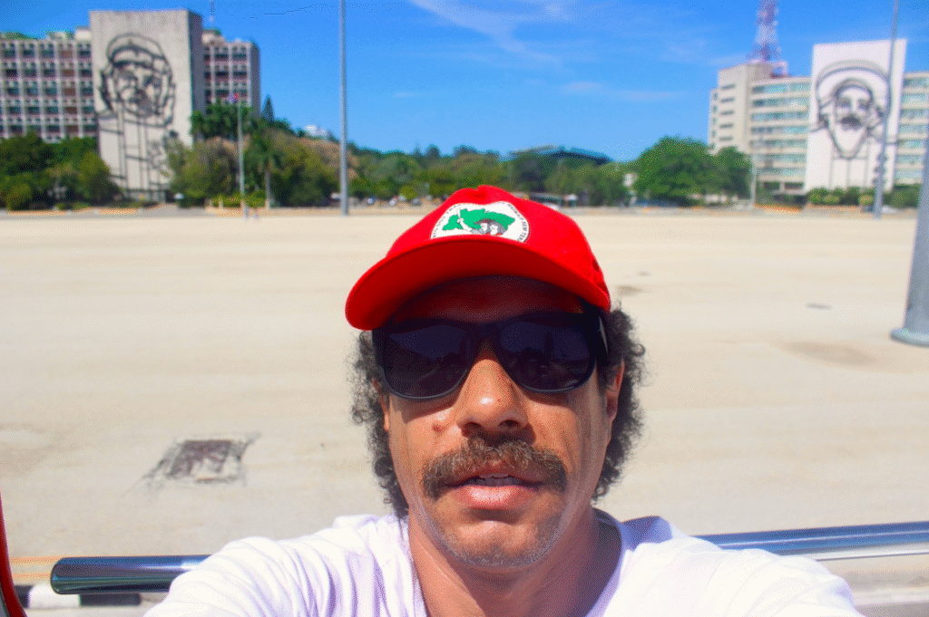 Tiago Prado na Praça da Revolução, Cuba, com boné do MST, reforçando o "vai pra cuba eu fui" e o compromisso ideológico.