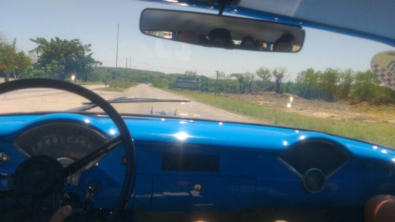 Vista de dentro de um carro clássico cubano azul, com a estrada e "Hasta la Victoria Siempre" ao fundo, mostrando a jornada e quanto custou ir para Cuba.