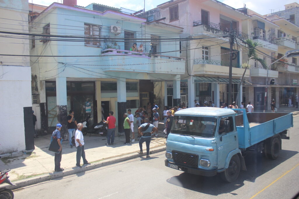 Pessoas em fila na calçada observando um caminhão azul antigo passar