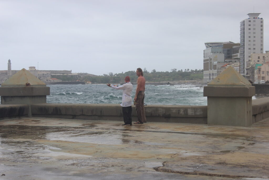 Pessoas no calçadão do Malecón observando o mar agitado com edifícios modernos ao fundo