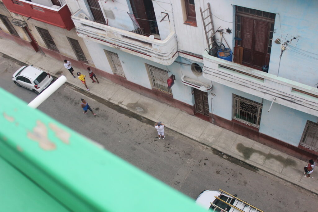 Vista de cima de uma rua em Havana, mostrando asfalto gasto, calçadas e fachadas de casas antigas.